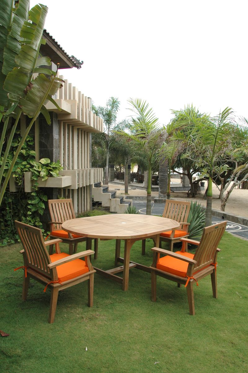 Outdoor wooden table with orange cushions surrounded by chairs on a grassy area with a building and trees in the background.