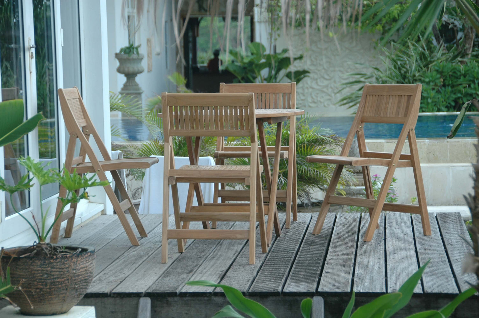 Wooden outdoor furniture set on a wooden deck with plants and a pool in the background
