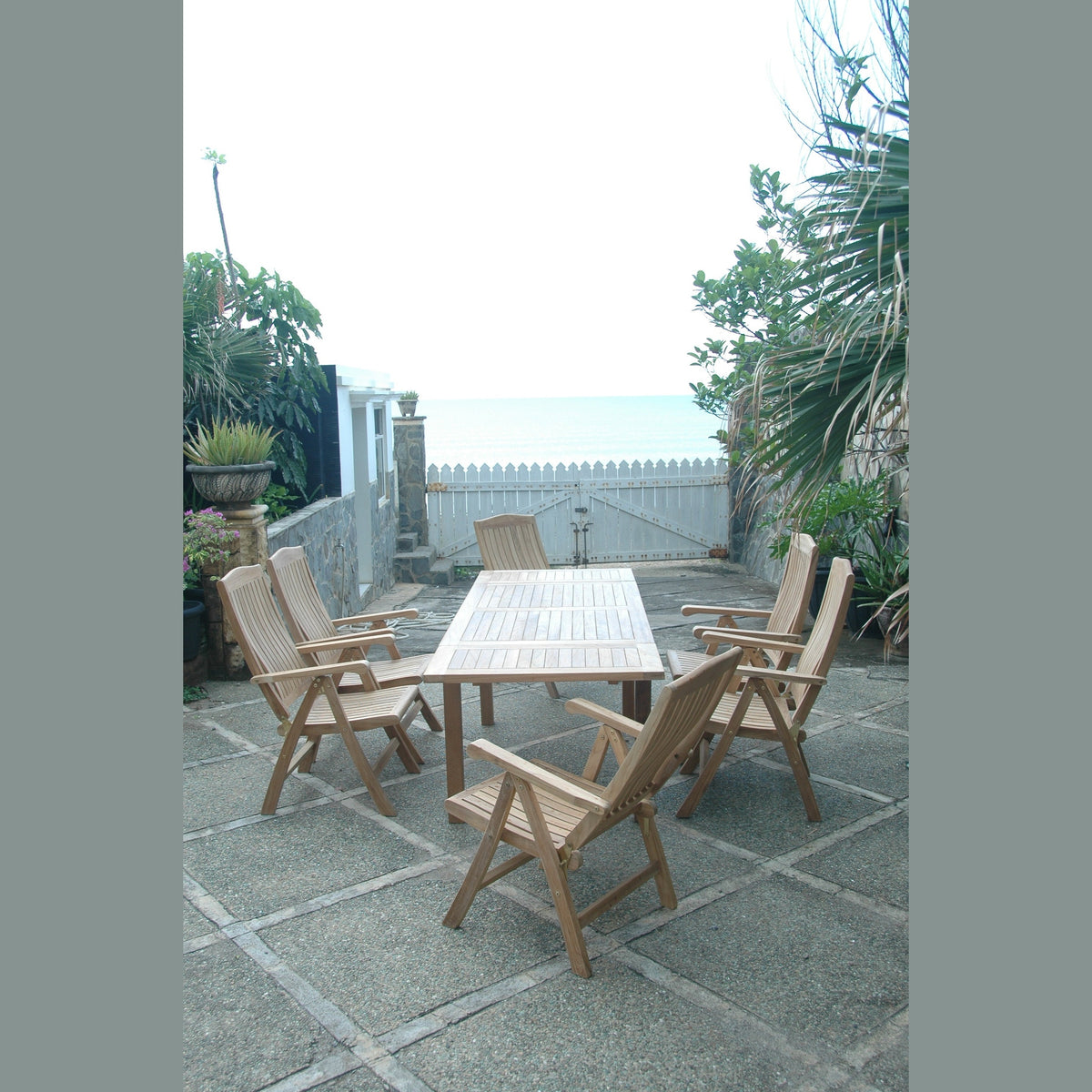 Wooden outdoor dining set on a stone patio with plants and a white fence in the background.