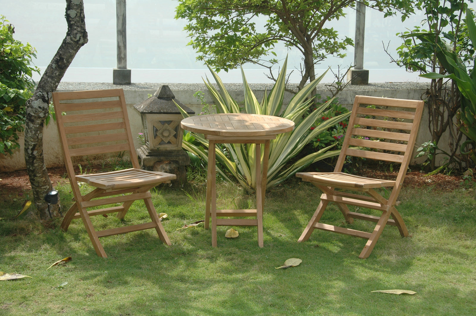 Two wooden chairs and a round table on a grassy area with plants and a wall in the background.