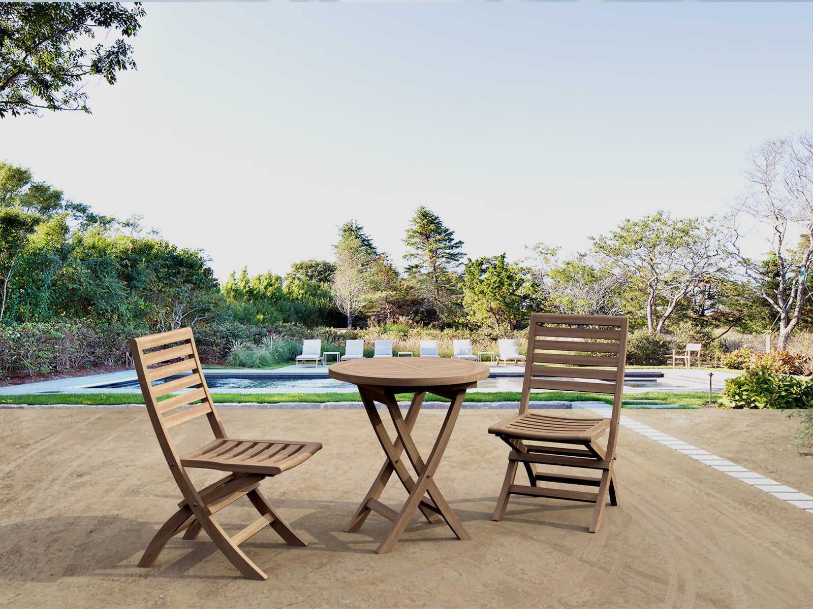 Wooden outdoor table with two chairs on a patio area with trees and a pool in the background.