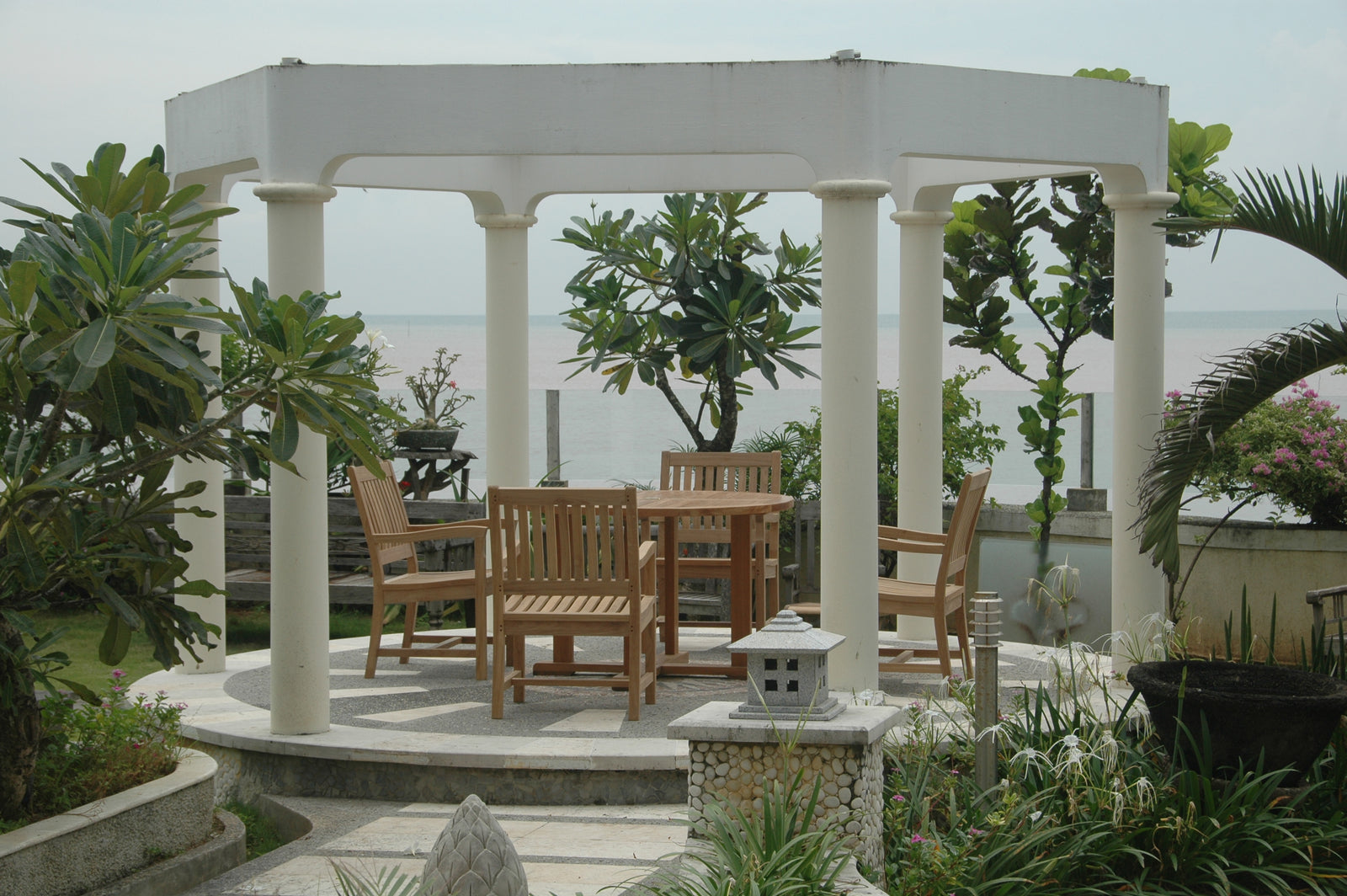 Patio with wooden furniture under a white pergola surrounded by greenery.