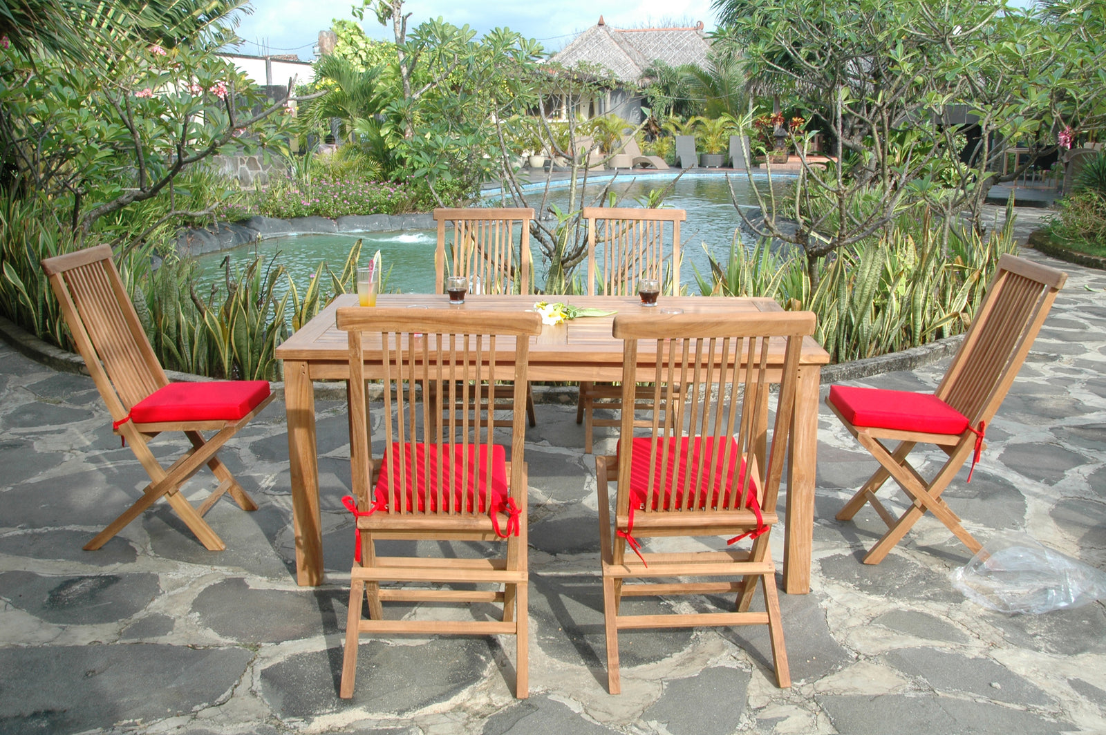 Outdoor wooden dining set with red cushions near a poolside garden.