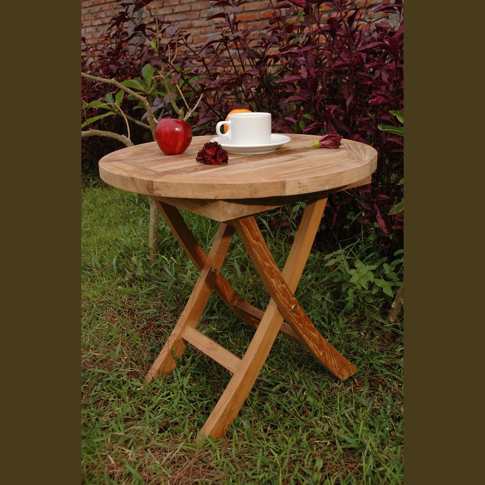 Wooden folding table with a cup, plate, and apples on a grassy area with plants in the background