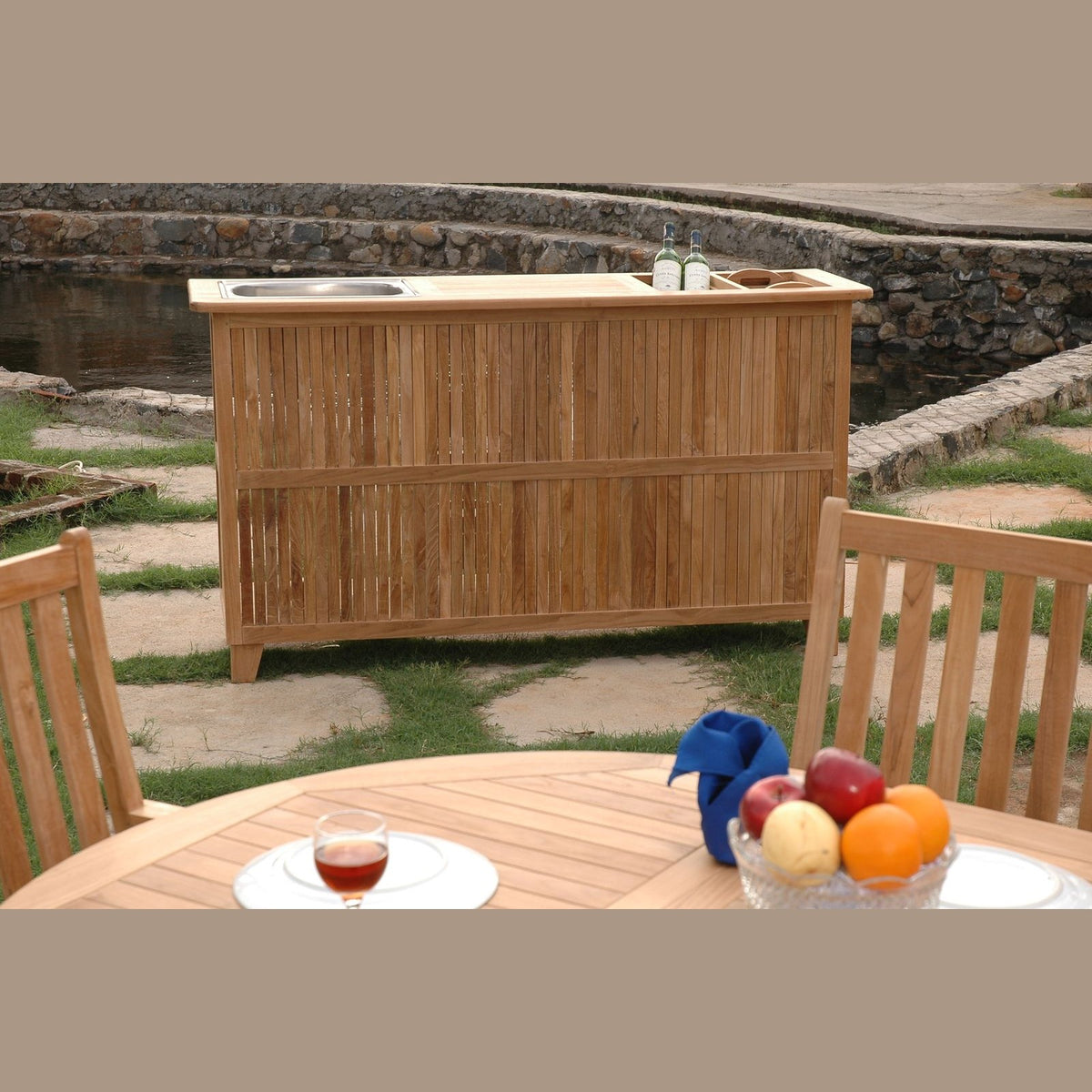 Wooden outdoor bar with stools and a table with fruit and drinks in a garden setting.