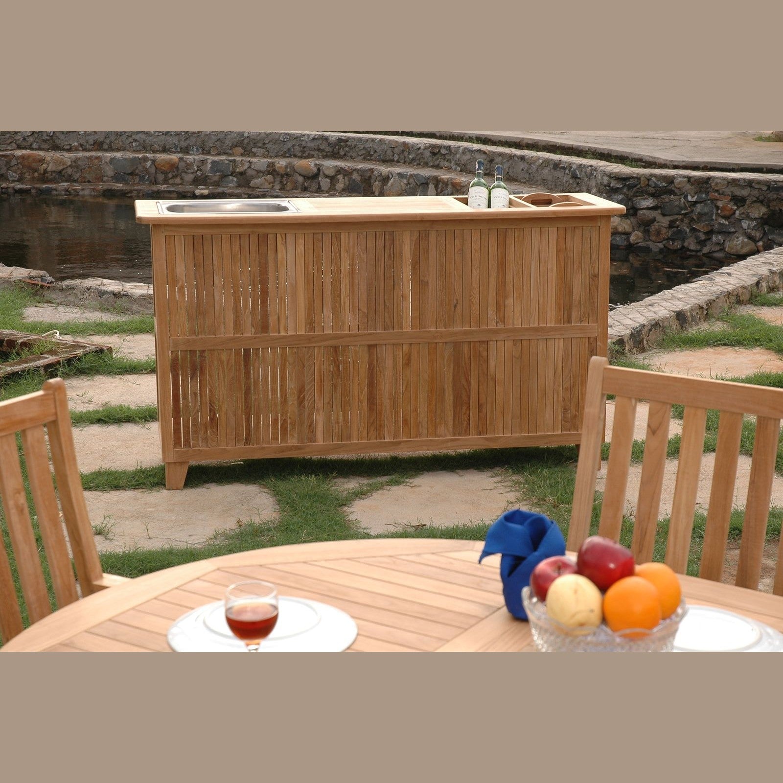 Wooden outdoor bar with stools and a table with fruit and drinks in a garden setting.