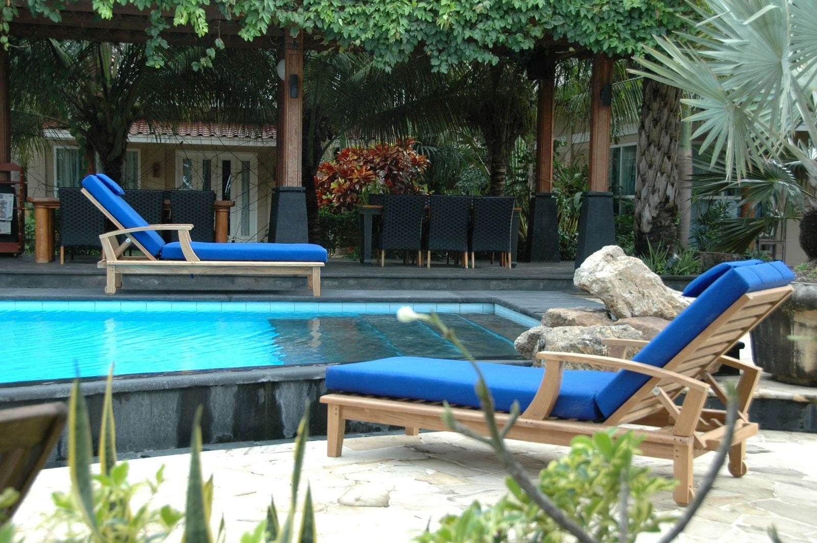 Two wooden lounge chairs with blue cushions by a poolside in a garden setting.