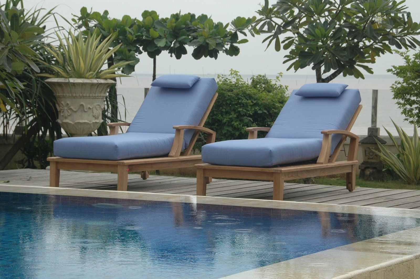 Two blue cushioned lounge chairs by a pool with greenery in the background