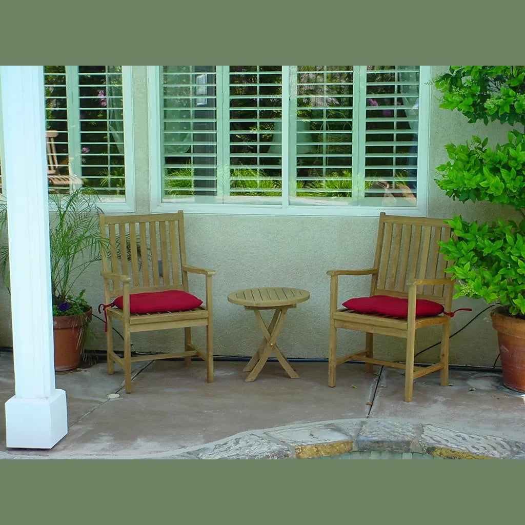Two wooden chairs with red cushions and a small round table on a patio with plants and a window in the background.