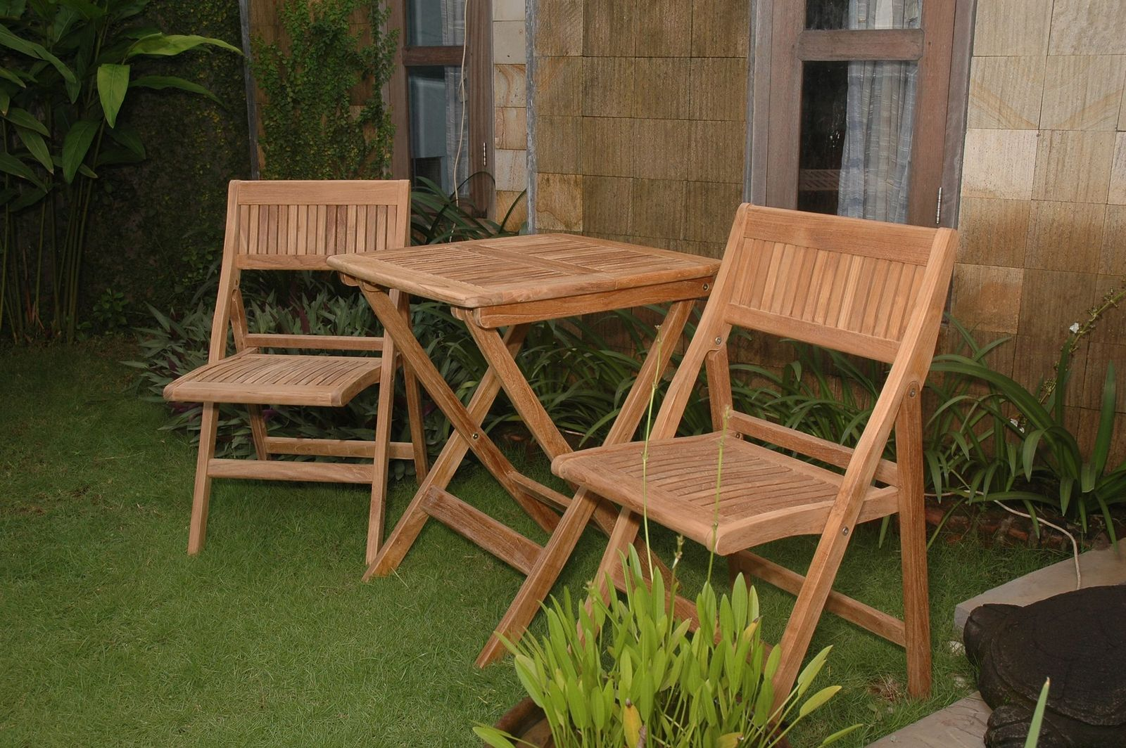 Wooden outdoor table and chairs on a grassy area with plants and a stone wall in the background.