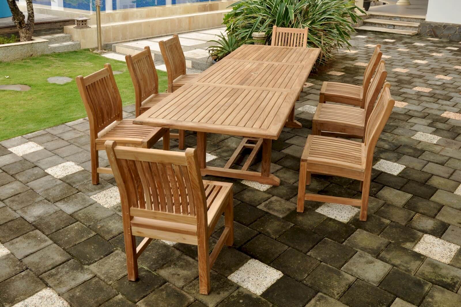 Wooden outdoor dining set on a paved patio with grass and plants in the background.