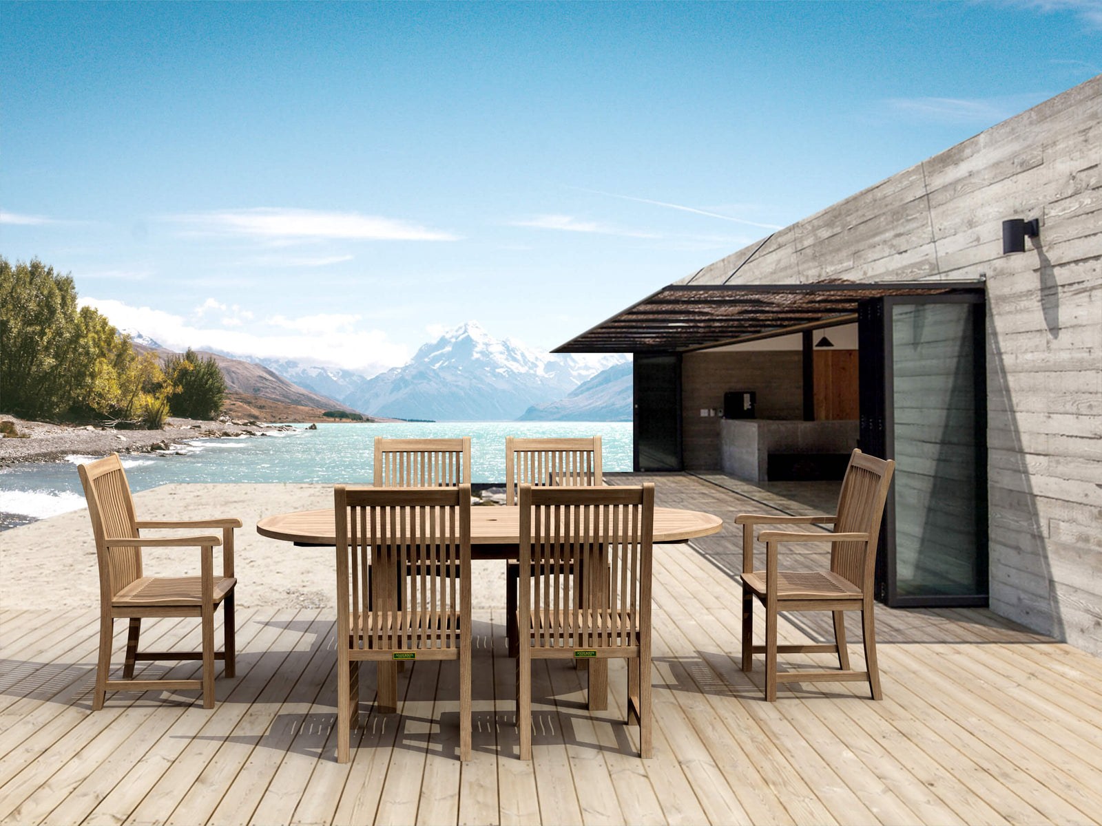Outdoor dining area with wooden table and chairs on a deck overlooking a scenic lake and mountains.