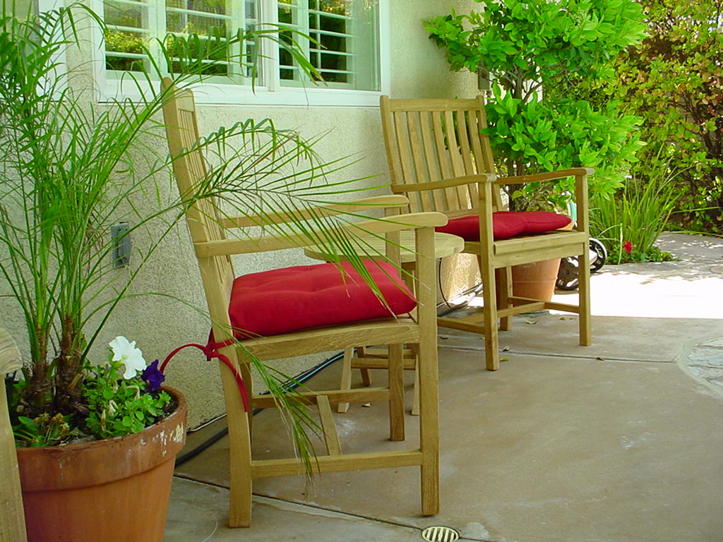 Two wooden chairs with red cushions on a patio, surrounded by plants.