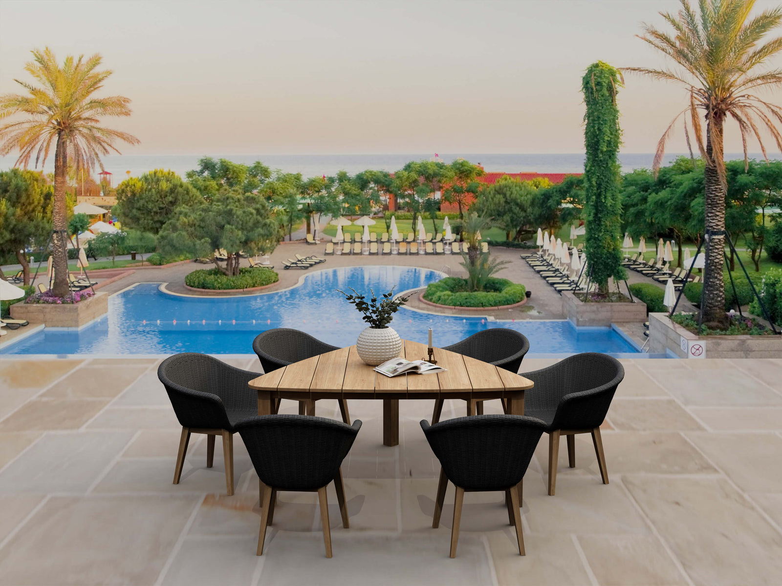 Outdoor dining area with a wooden table and black chairs near a pool with palm trees and greenery.