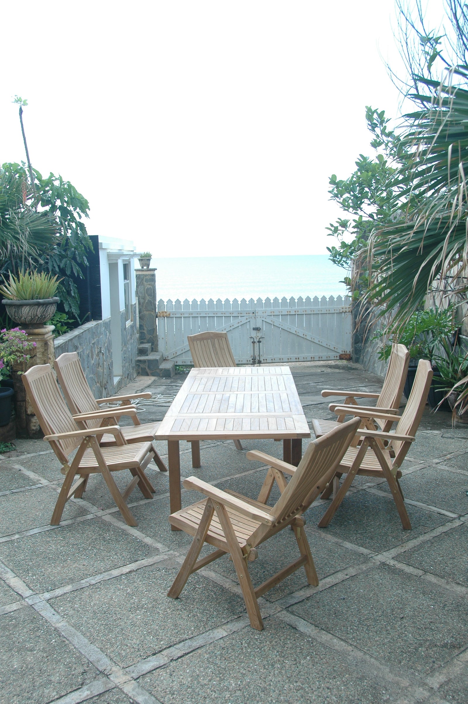 Wooden outdoor dining set on a stone patio with plants and a white fence in the background.