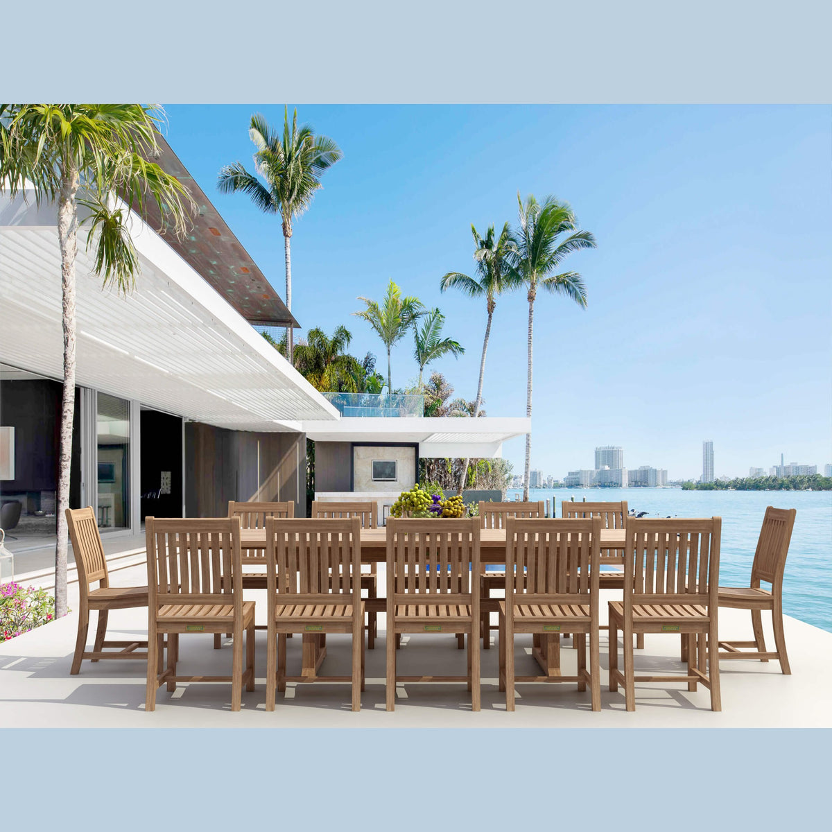 Outdoor dining set with wooden chairs and table by a waterfront with palm trees and a house in the background.