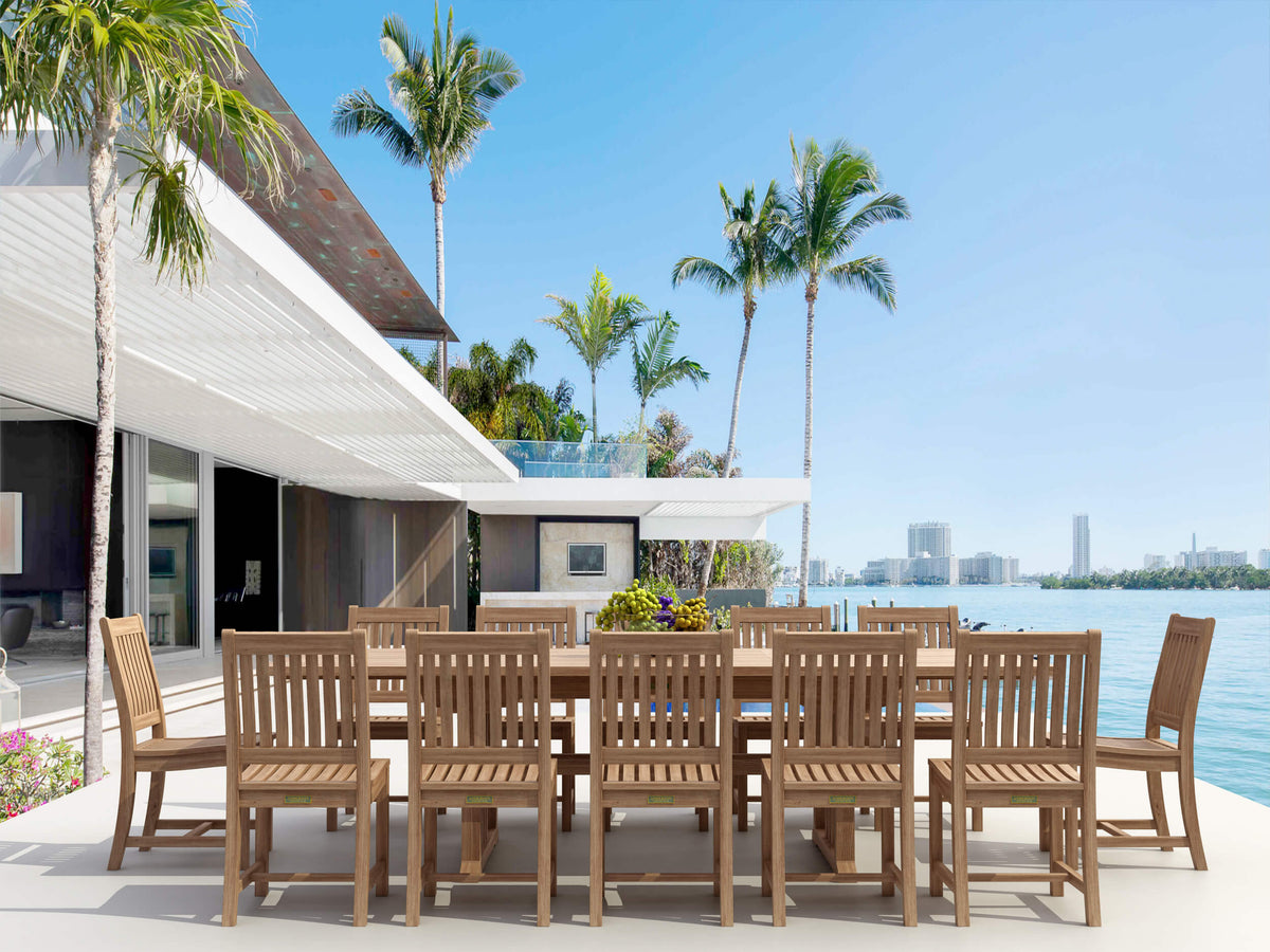 Outdoor dining set with wooden chairs and table by a waterfront with palm trees and a house in the background.