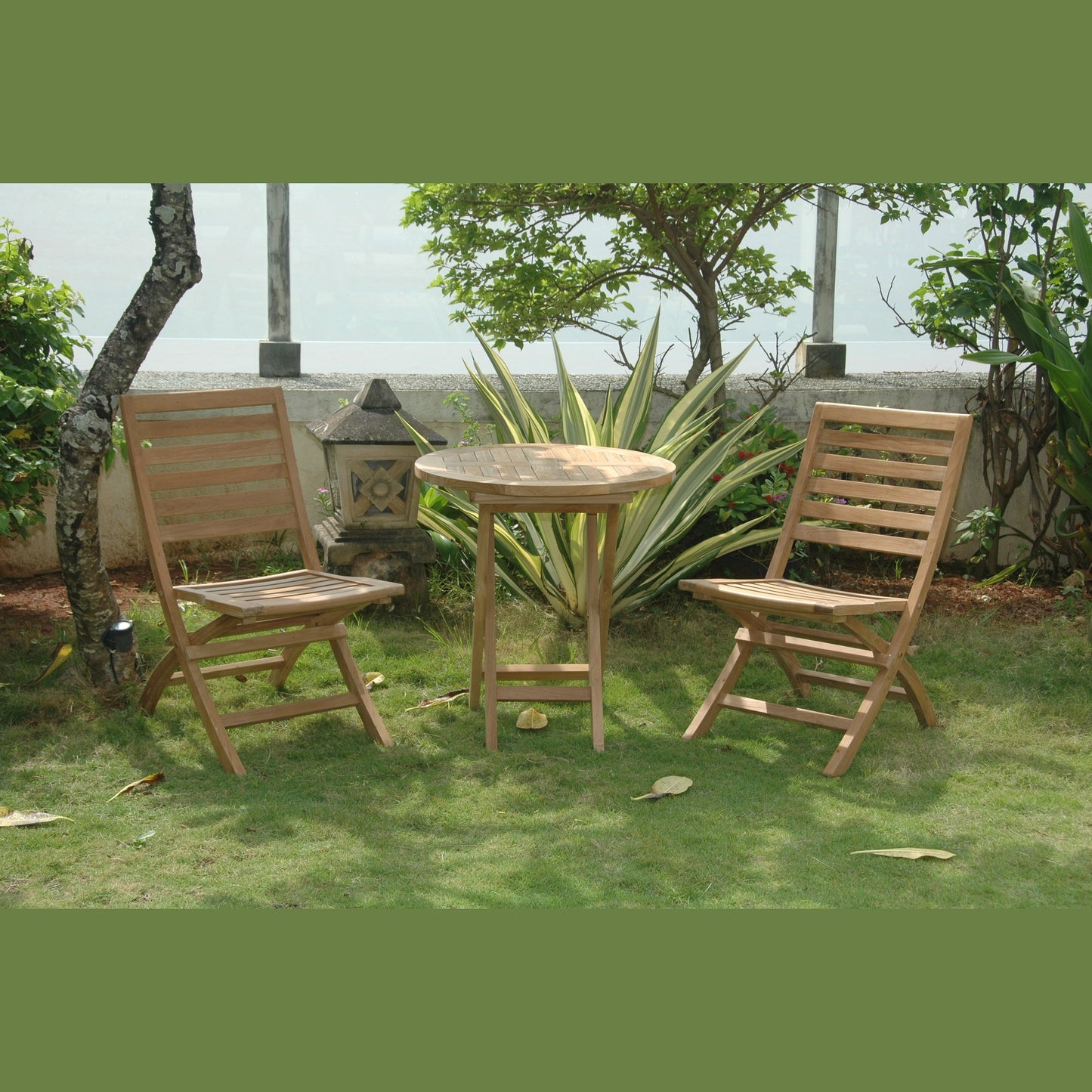 Two wooden chairs and a round table on a grassy area with plants and a wall in the background.