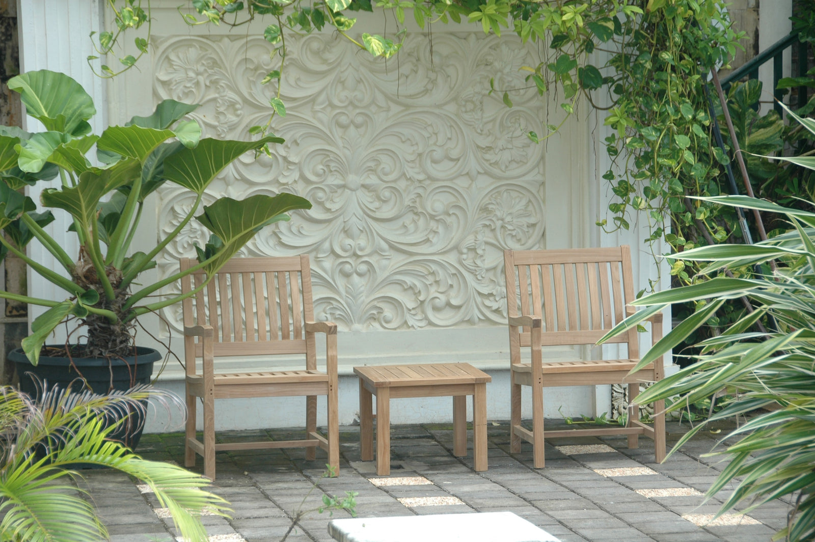Two wooden chairs and a small table on a patio with decorative wall and plants.
