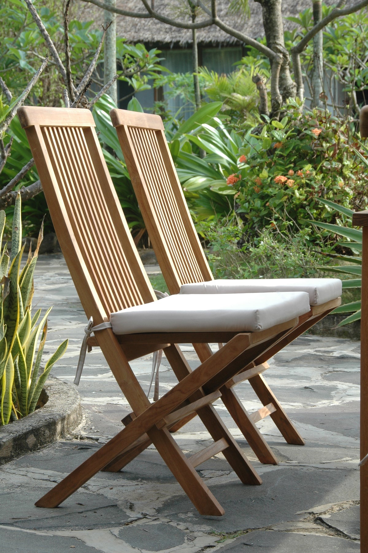 Two wooden folding chairs with white cushions on a stone patio surrounded by greenery.