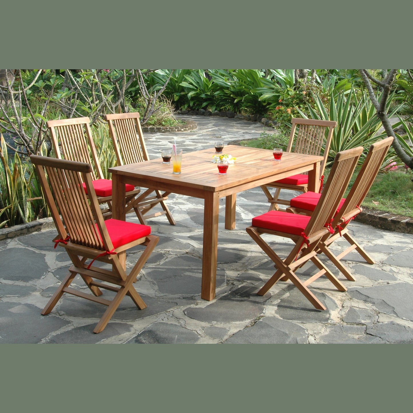 Wooden outdoor dining table with six chairs on a stone patio surrounded by greenery.