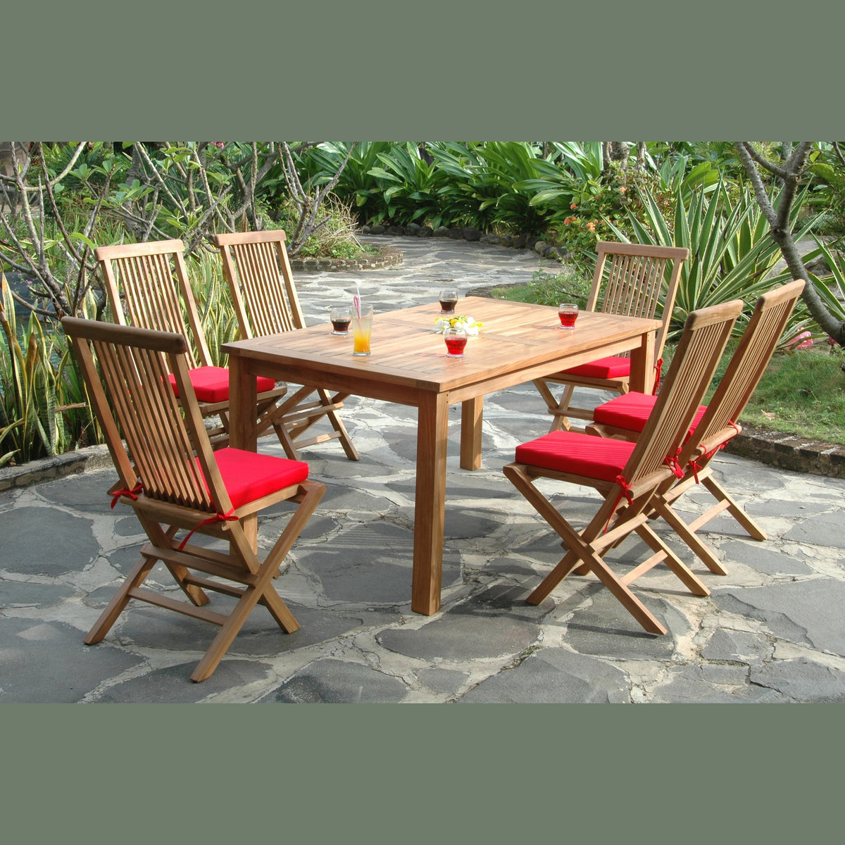 Wooden outdoor dining table with six chairs on a stone patio surrounded by greenery.