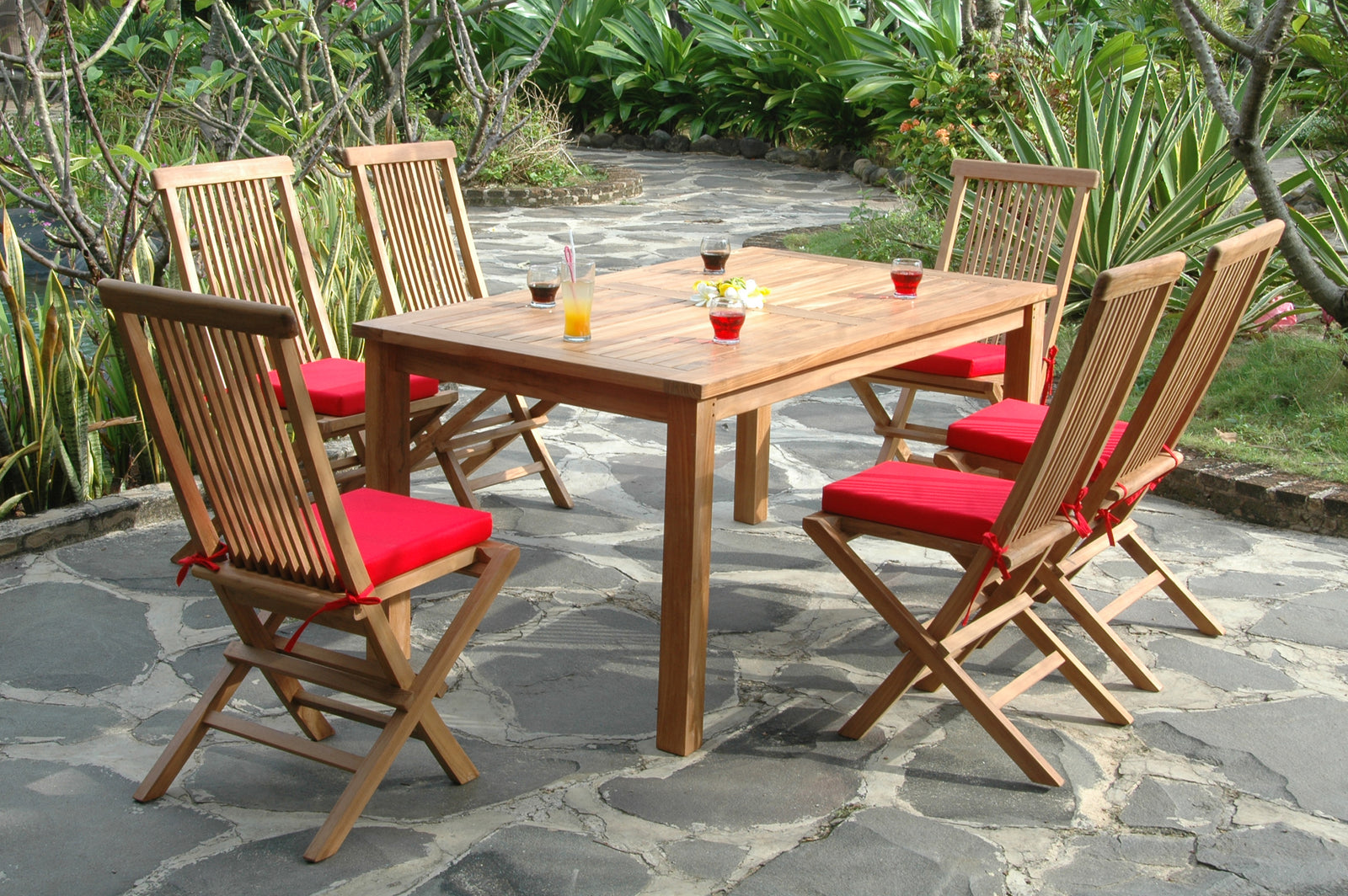 Wooden outdoor dining table with six chairs on a stone patio surrounded by greenery.