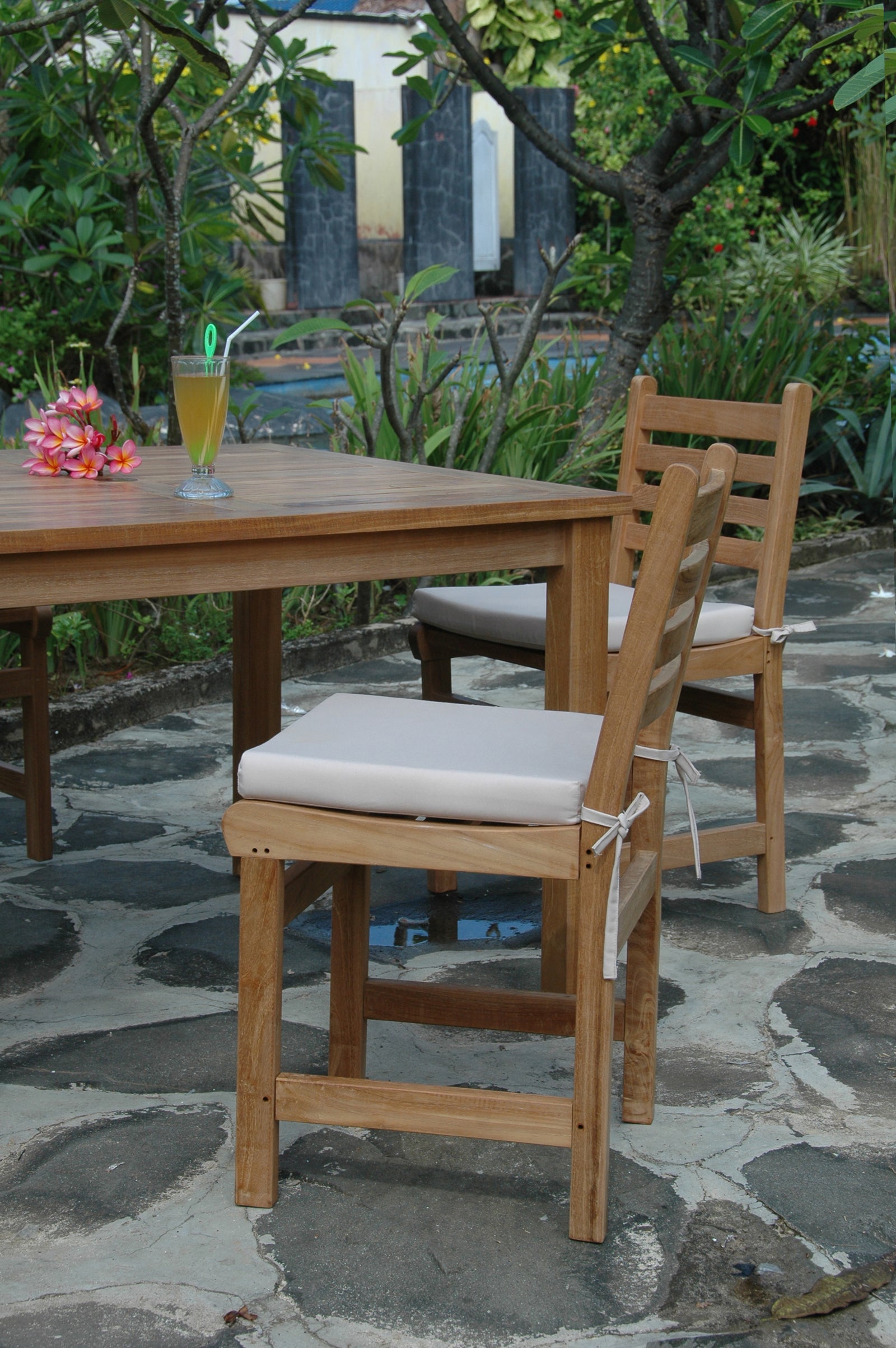 Wooden chairs with white cushions on a stone patio with a table and drink in the background.