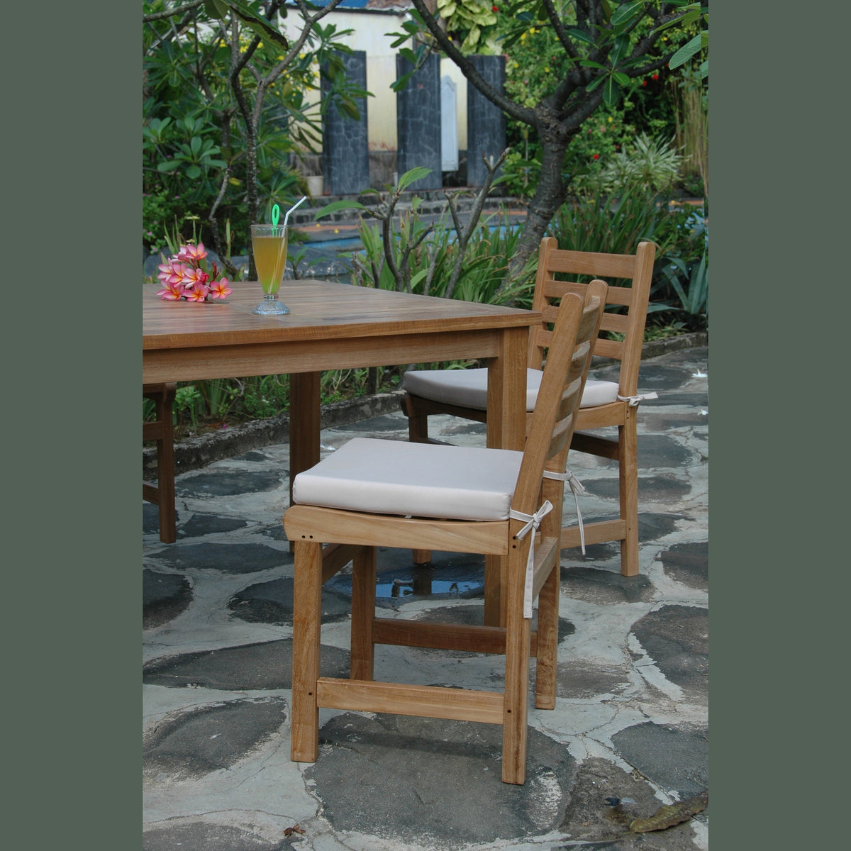 Wooden chairs with white cushions on a stone patio with a table and drink in the background.