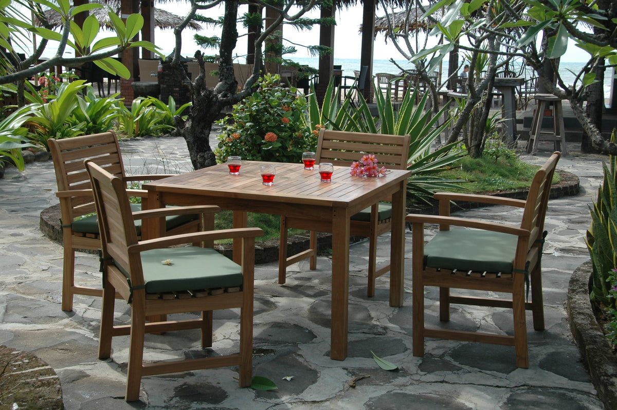 Outdoor patio set with wooden table and chairs on a stone patio surrounded by greenery.