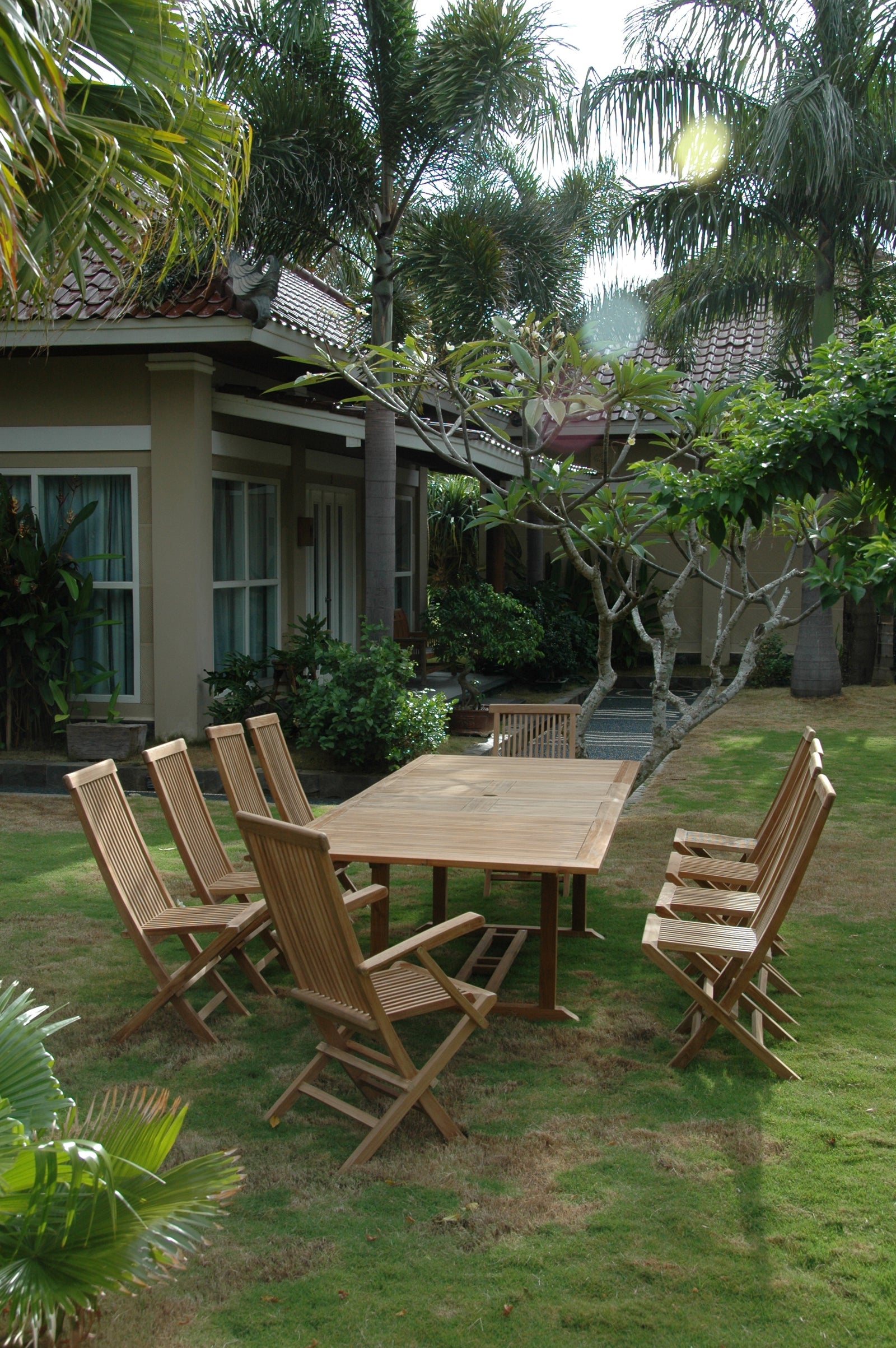 Wooden outdoor dining set with chairs on a grassy area surrounded by trees and a building.