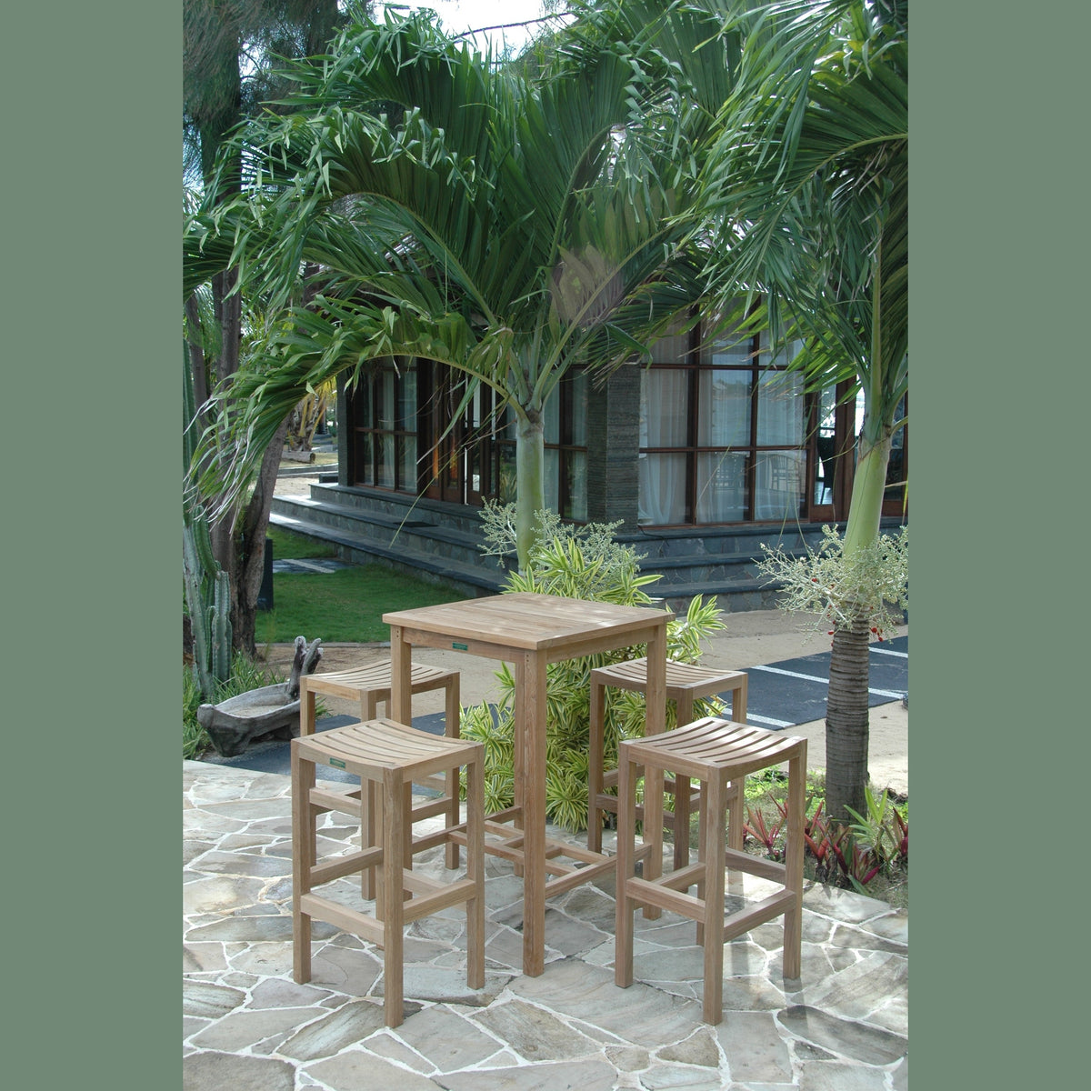 Outdoor setting with wooden table and stools surrounded by palm trees and a building in the background.