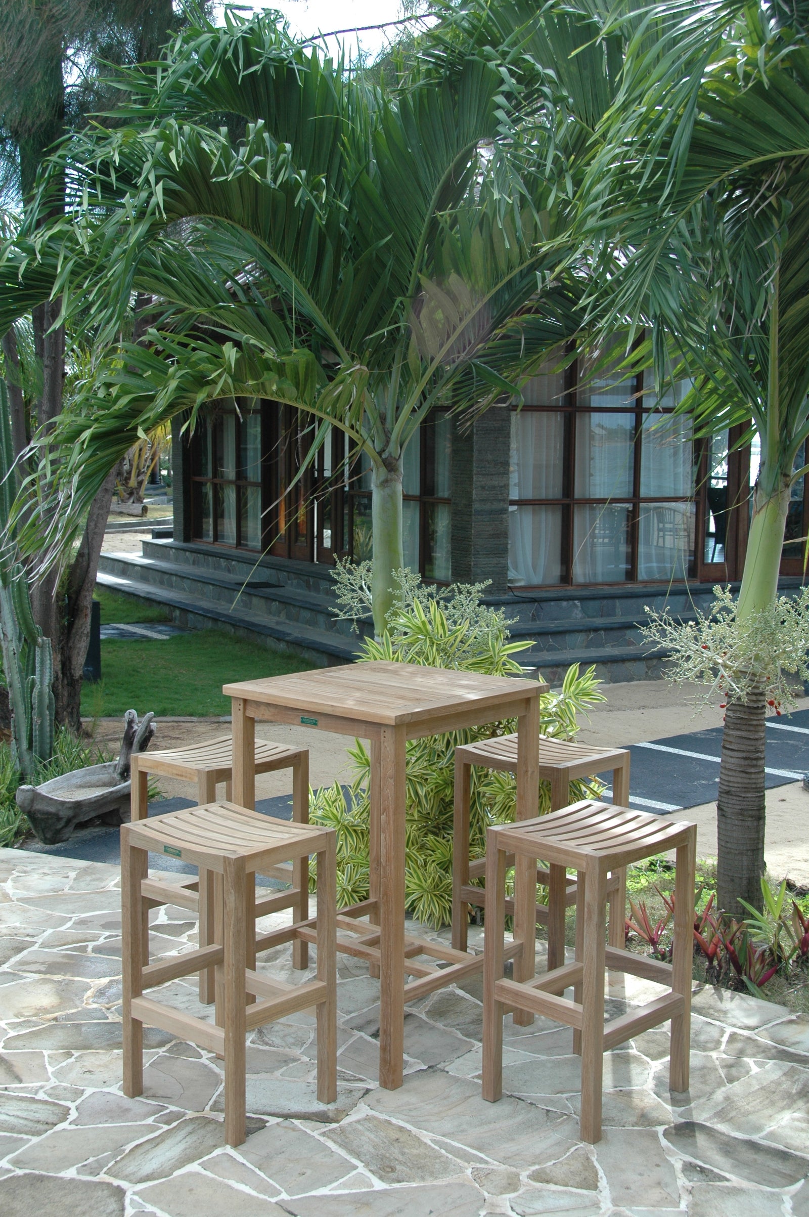 Outdoor setting with wooden table and stools surrounded by palm trees and a building in the background.