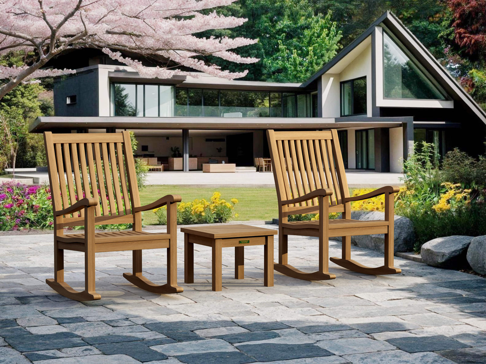 Two wooden rocking chairs and a small table on a patio in front of a modern house with cherry blossoms.
