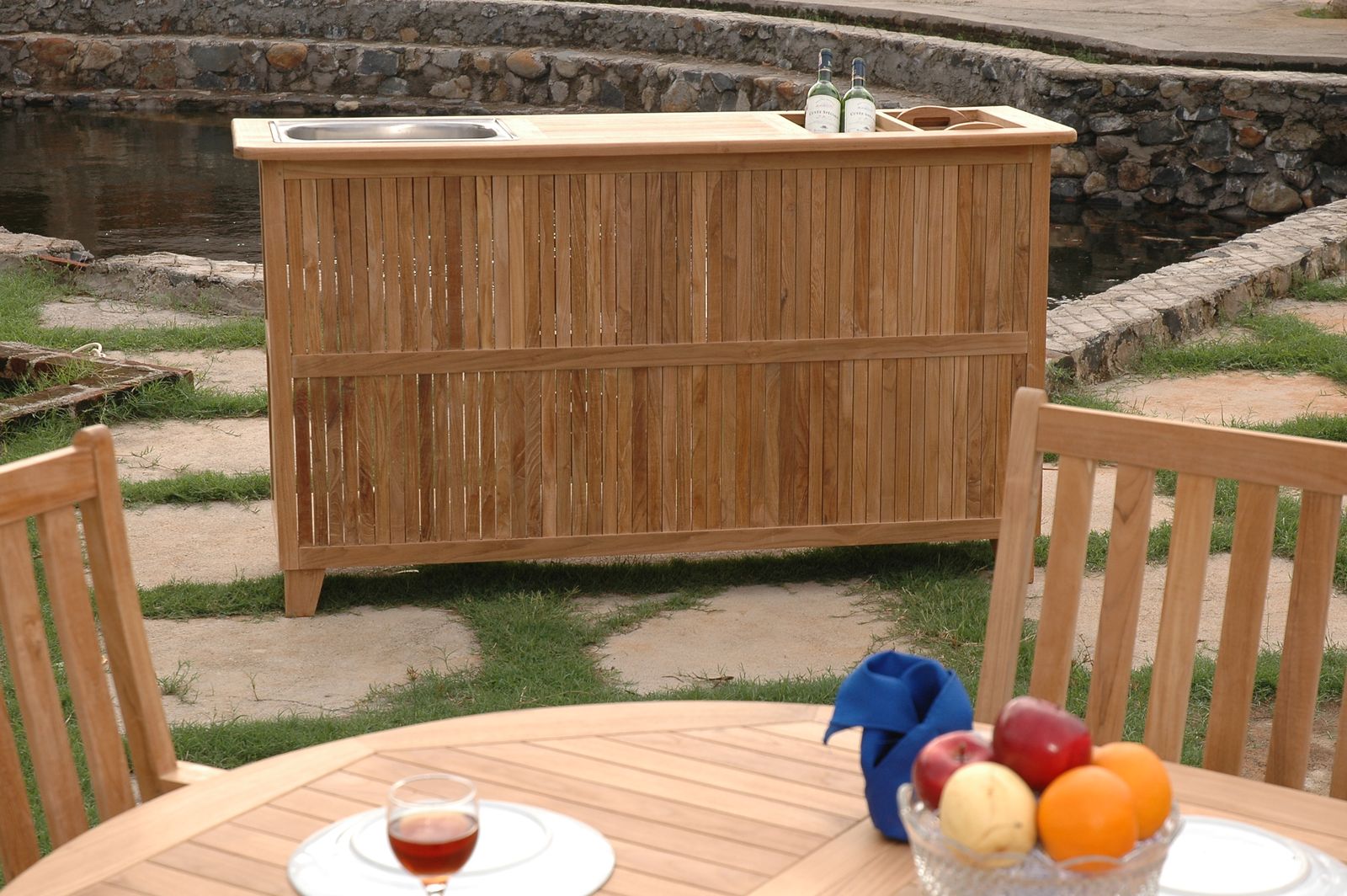 Wooden outdoor bar with stools and a table with fruit and drinks in a garden setting.