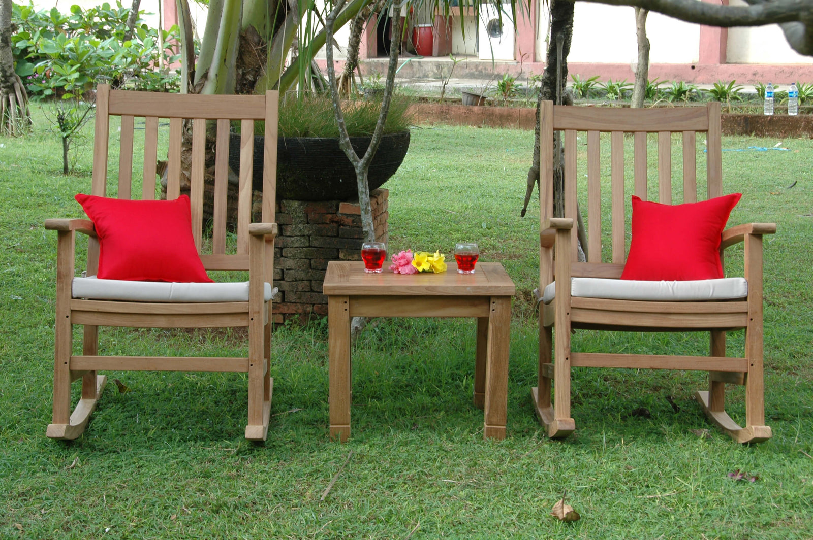 Two wooden rocking chairs with red cushions and a small table in a garden setting.