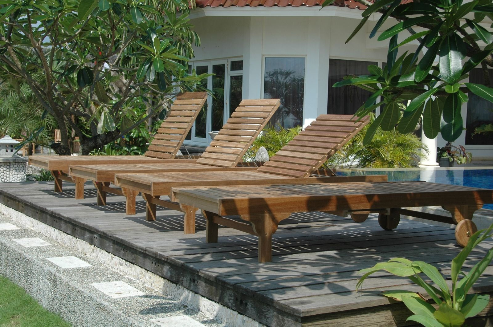 Row of wooden lounge chairs by a poolside with a building and trees in the background.