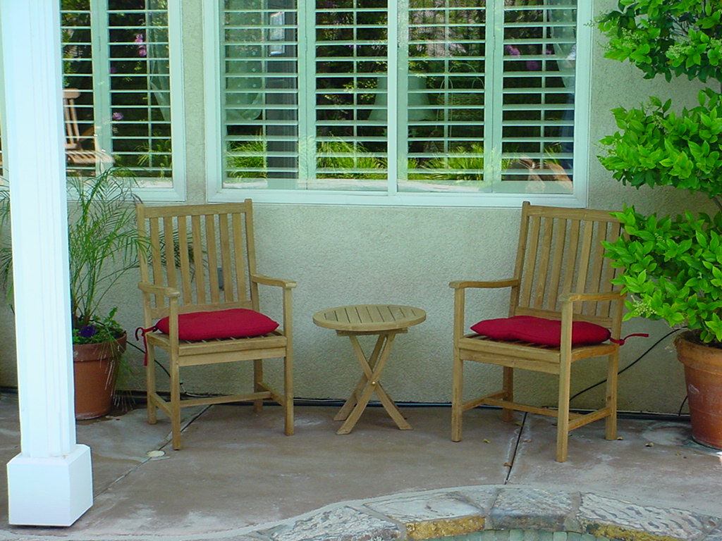 Two wooden chairs with red cushions and a small round table on a patio with plants and a window in the background.