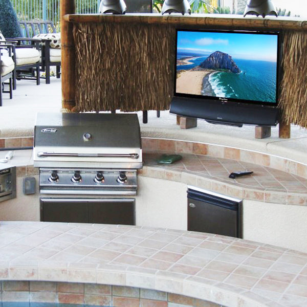 Outdoor kitchen with grill, refrigerator, and TV displaying a beach scene.