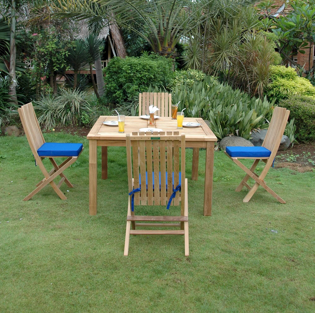 Wooden outdoor table with four chairs on a grassy area with greenery in the background