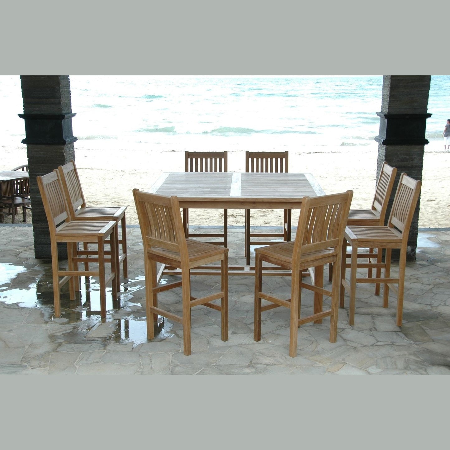 Wooden outdoor dining set with table and chairs on a stone patio by the beach.