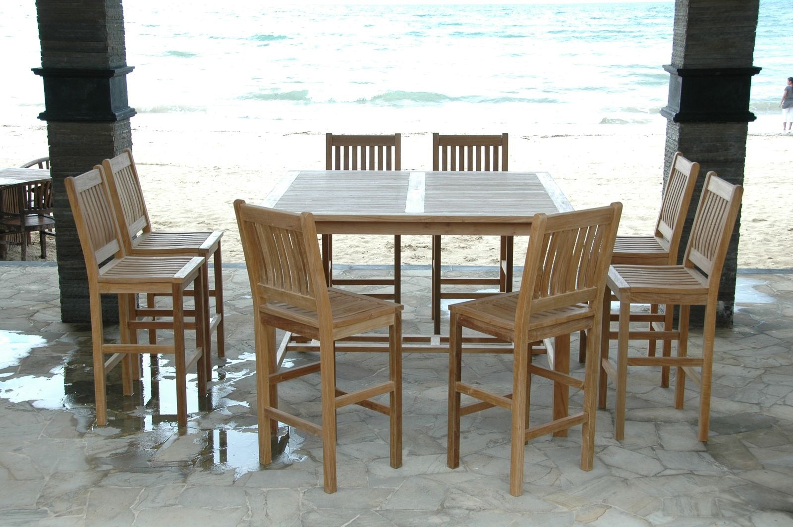 Wooden outdoor dining set with table and chairs on a stone patio by the beach.