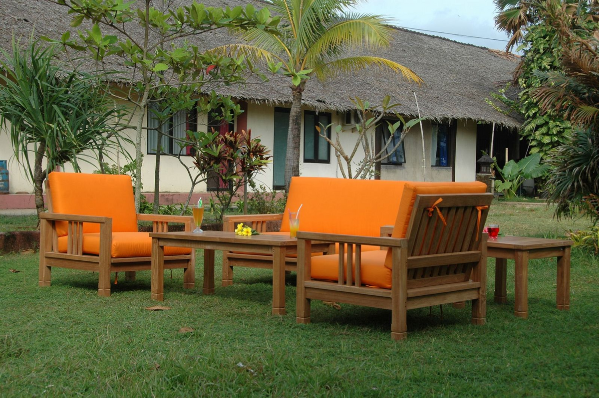 Outdoor furniture set with orange cushions on a grassy area in front of a thatched-roof building.