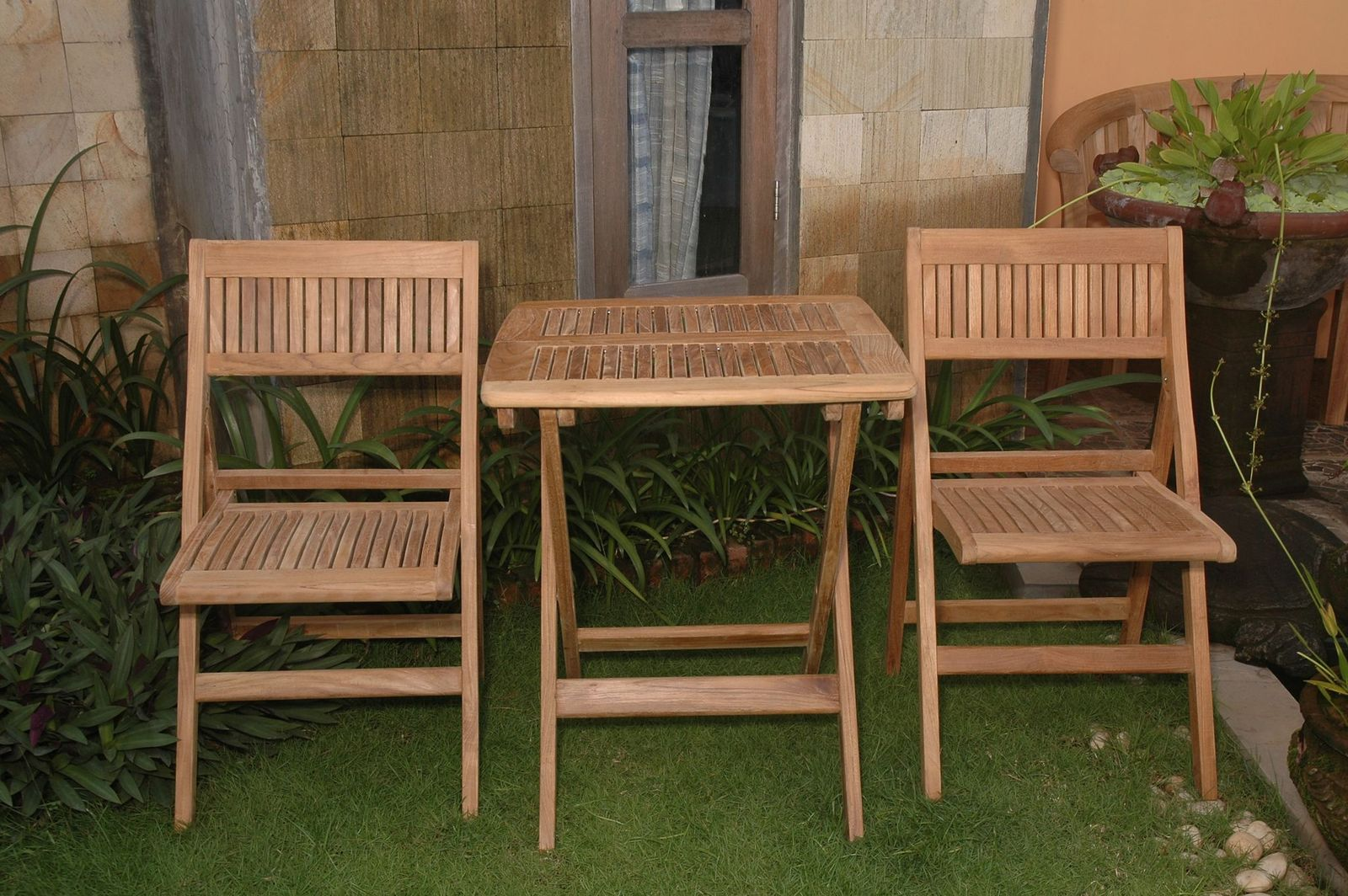 Wooden outdoor table and chairs set on a grassy area with plants and a stone wall in the background.