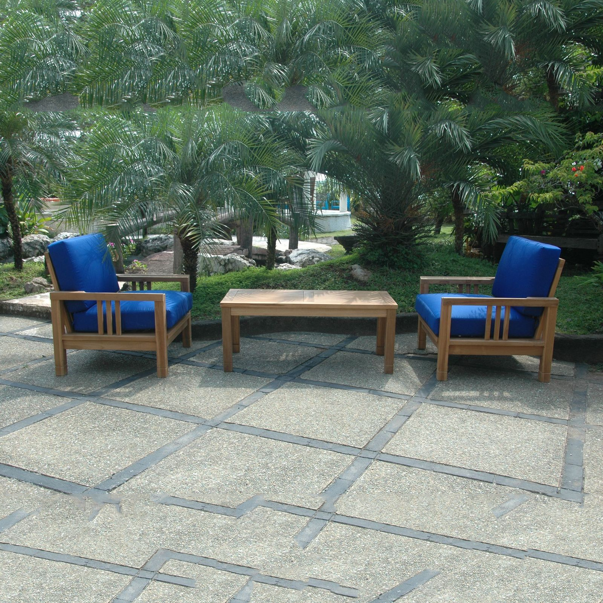 Two wooden chairs with blue cushions and a wooden table on a stone patio with greenery in the background.